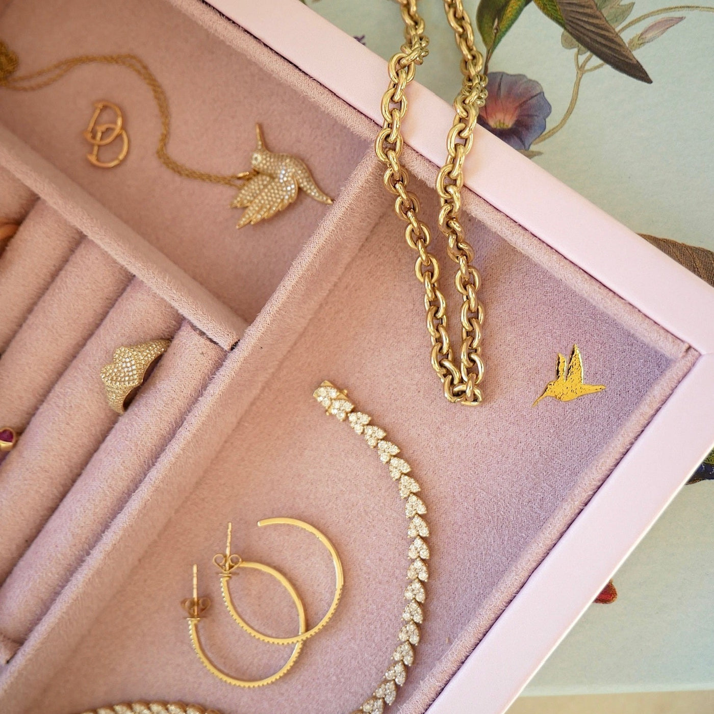 Signature Jewelry Box in the color pink embossed in corner with a golden hummingbird, displayed with 14k yellow gold jewelry with diamonds