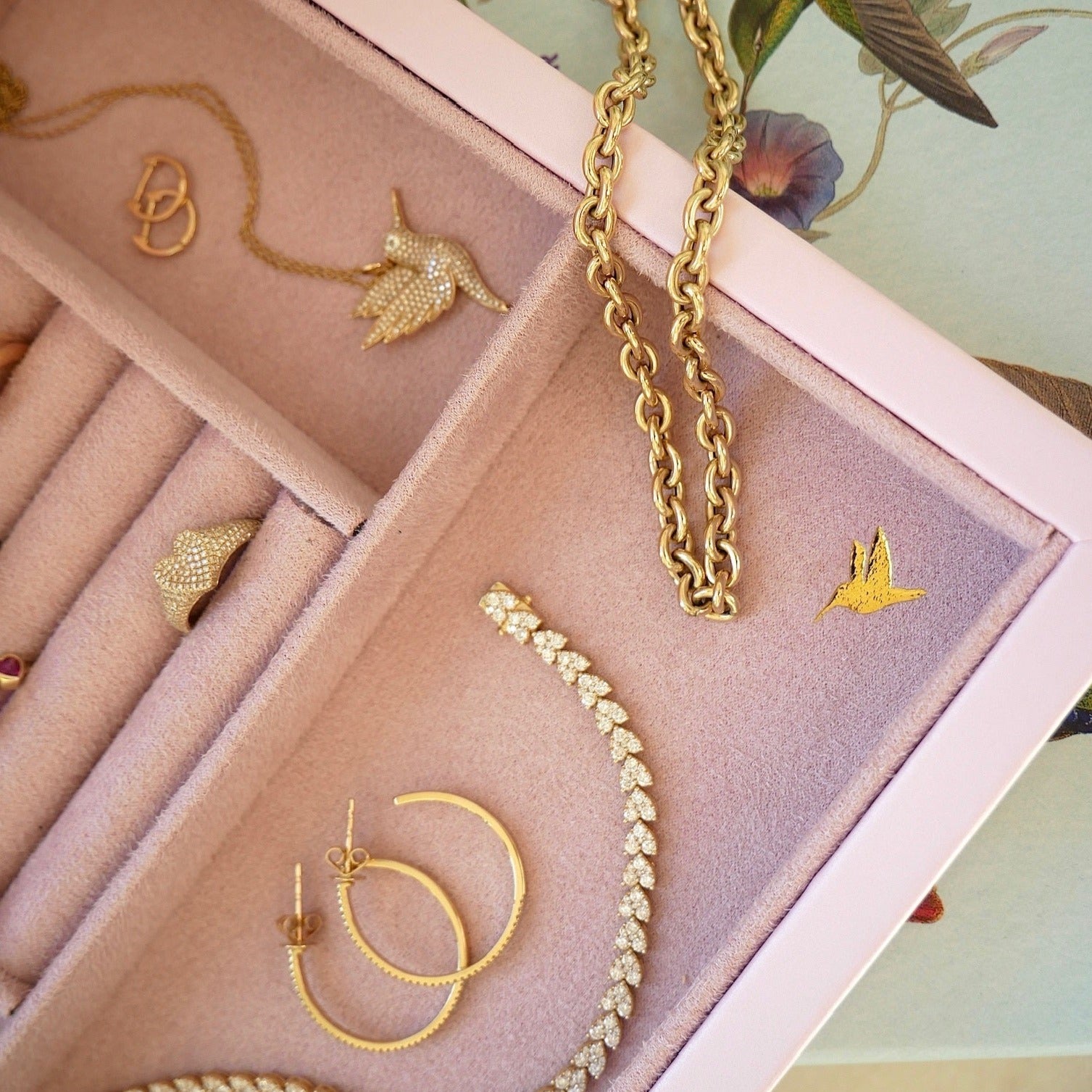 Signature Jewelry Box in the color pink embossed in corner with a golden hummingbird, displayed with 14k yellow gold jewelry with diamonds
