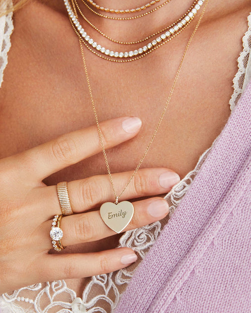 Gold Jumbo Heart Necklace in 14k yellow gold engraved with initials Emily, styled on neck of model with gold and diamond necklaces and rings