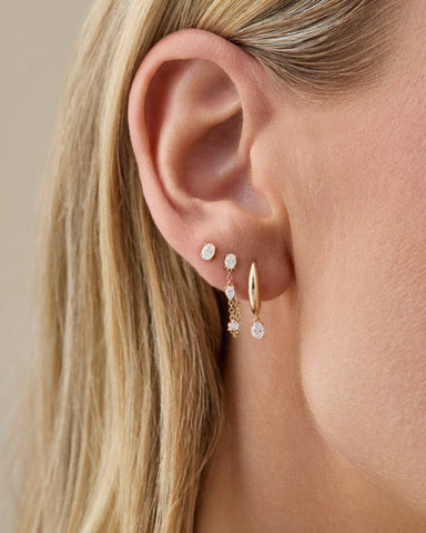 Close-up of an ear wearing gold hoop earrings with small gemstones against a neutral background