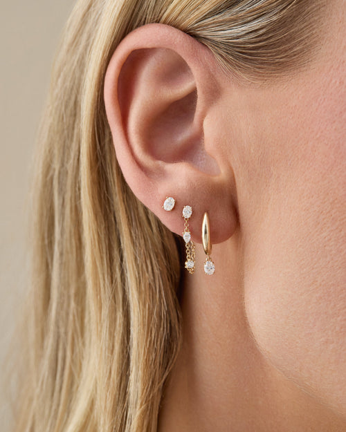 Close-up of an ear wearing gold hoop earrings with small gemstones against a neutral background