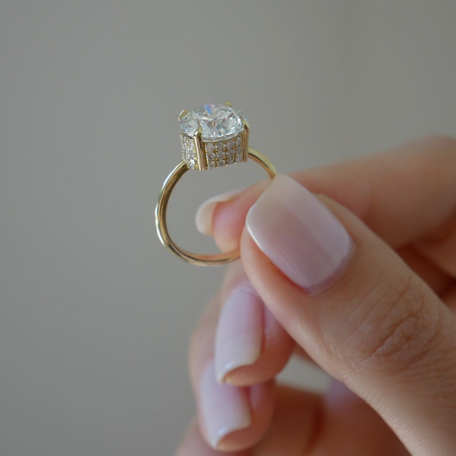 The Lindsay Engagement Ring with round center diamond, basket covered in pave diamonds, and yellow gold band held in between fingers of model