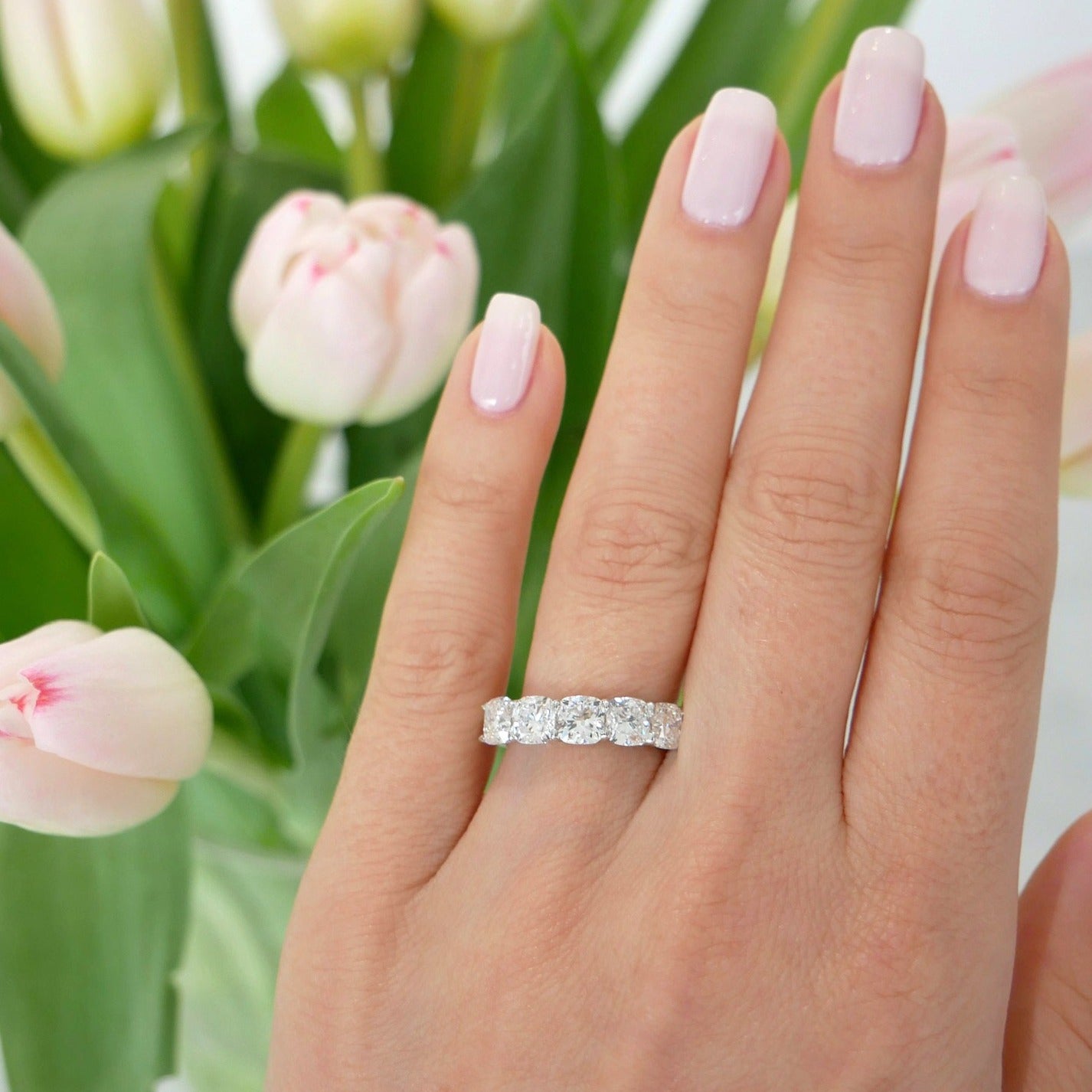 Oval Diamond Eternity Band in platinum setting styled on ring finger of model next to pink tulips 