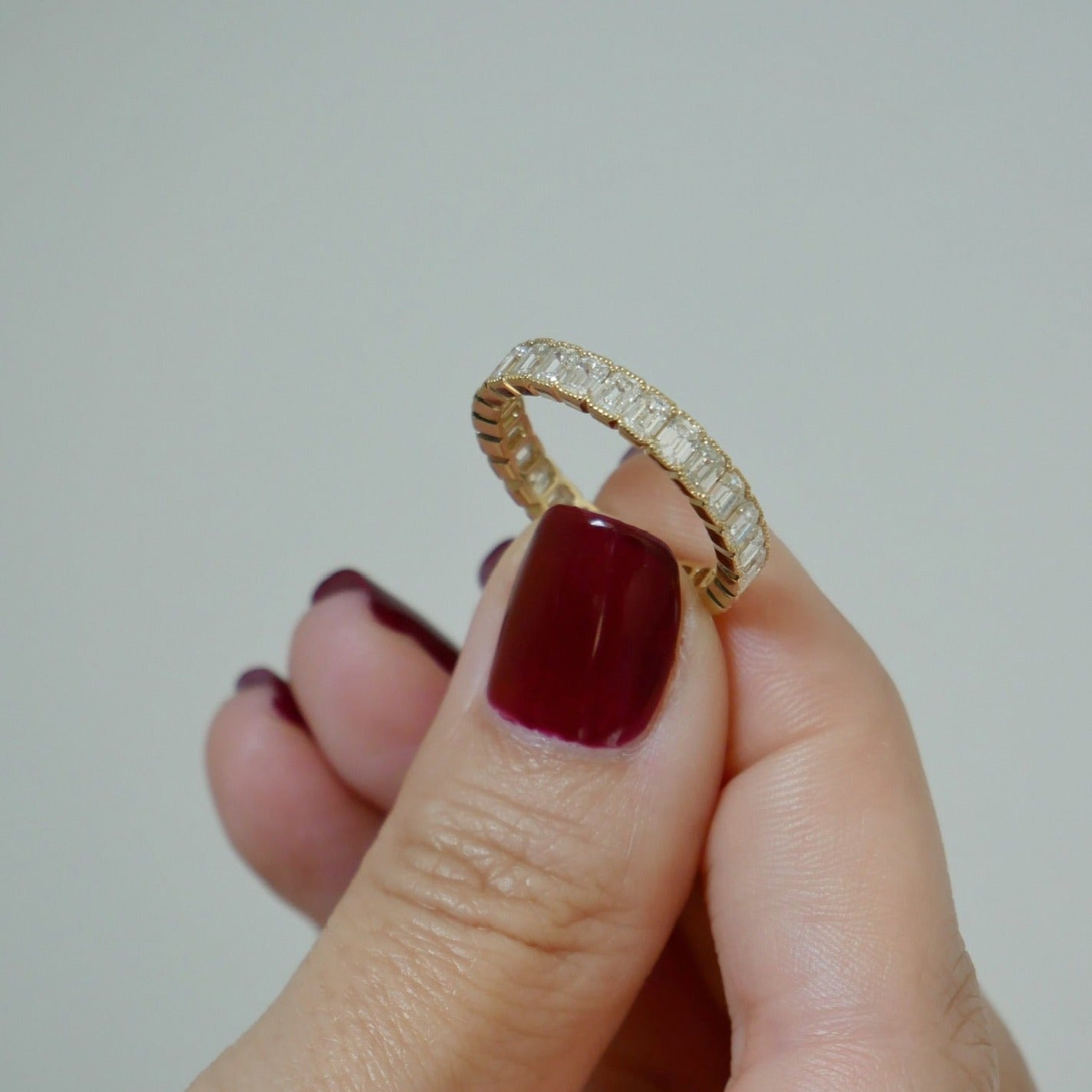 Emerald Cut Beaded Bezel Eternity Band in 18k yellow gold held in between fingers of model