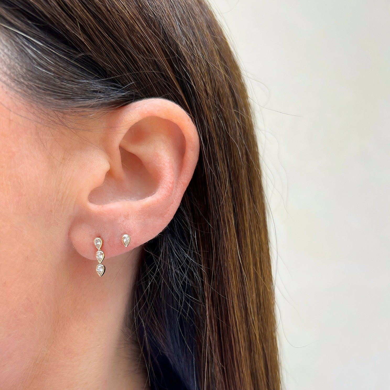 Bezel Set Triple Diamond Pear Stud Earring in 14k yellow gold next to diamond stud styled on ear lobe of model