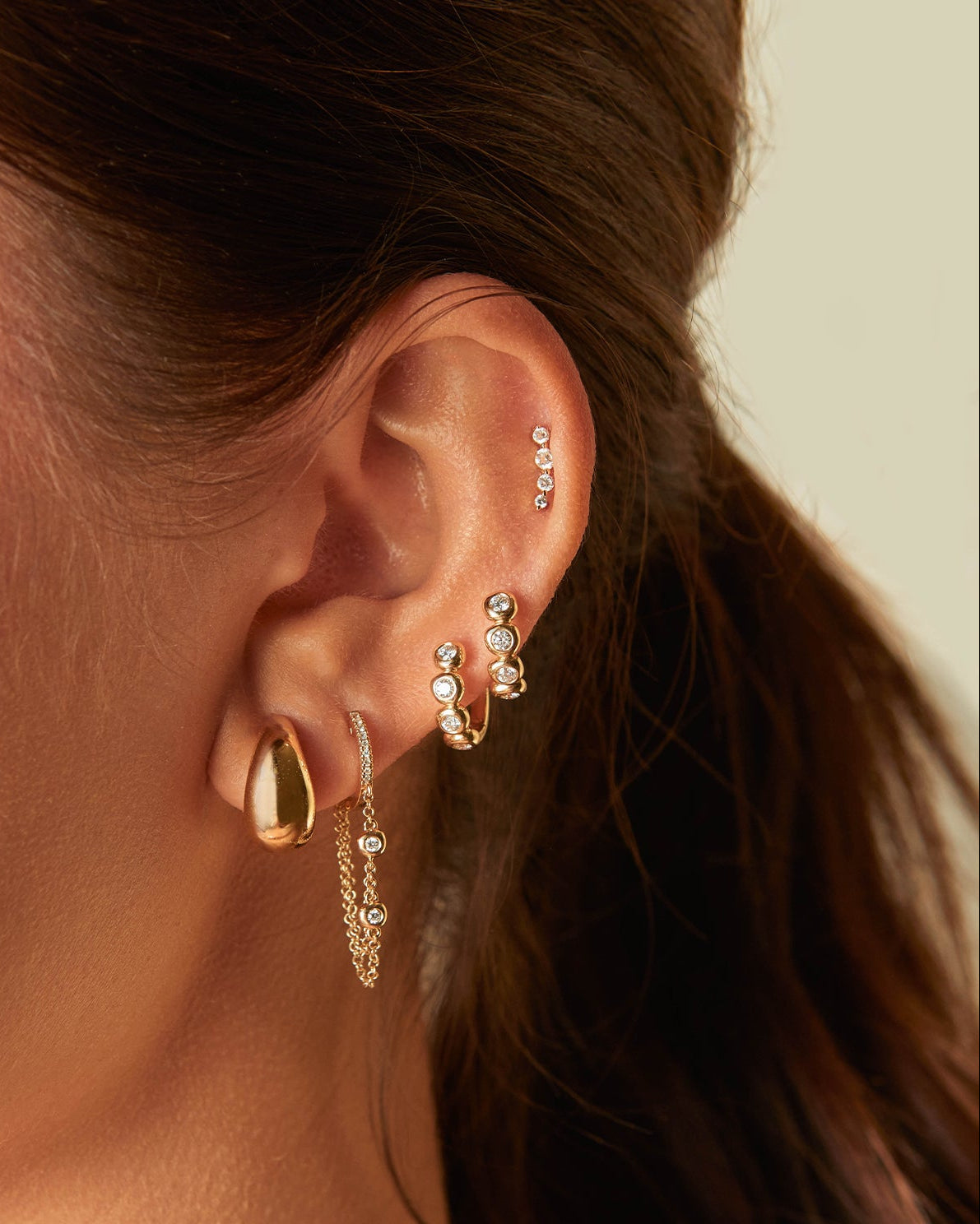 Prong Set Diamond Arc Stud Earring in 14k yellow gold styled on cartilage of ear of model next to four diamond and gold earrings 