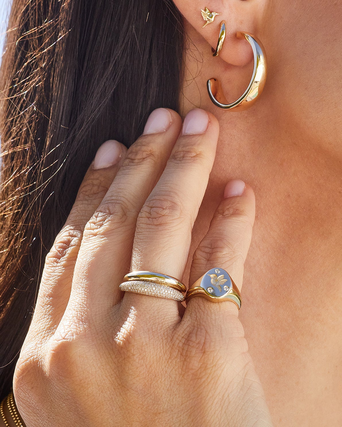 Diamond Hummingbird Signet Ring in 14k yellow gold styled on pinky ring of model next to diamond and gold dome ring styled on ring finger and gold earrings styled on ear of model