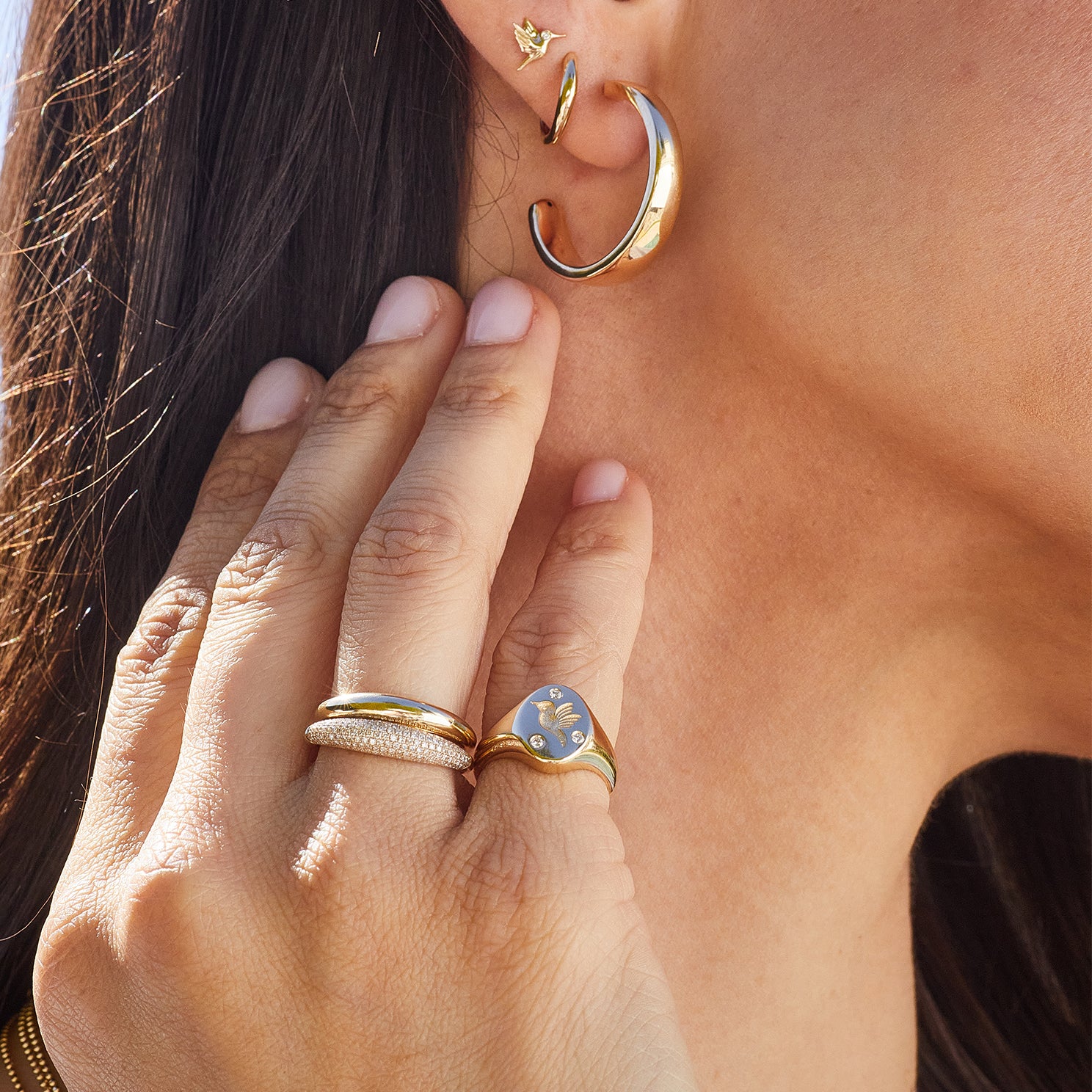 Diamond Hummingbird Signet Ring in 14k yellow gold styled on pinky ring of model next to diamond and gold dome ring styled on ring finger and gold earrings styled on ear of model