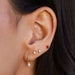 Full Cut Diamond Mini Teardrop Stud Earring in 14k yellow gold styled on second and third earring hole on ear lobe of model next to diamond mini teardrop huggie and ruby baguette stud earring