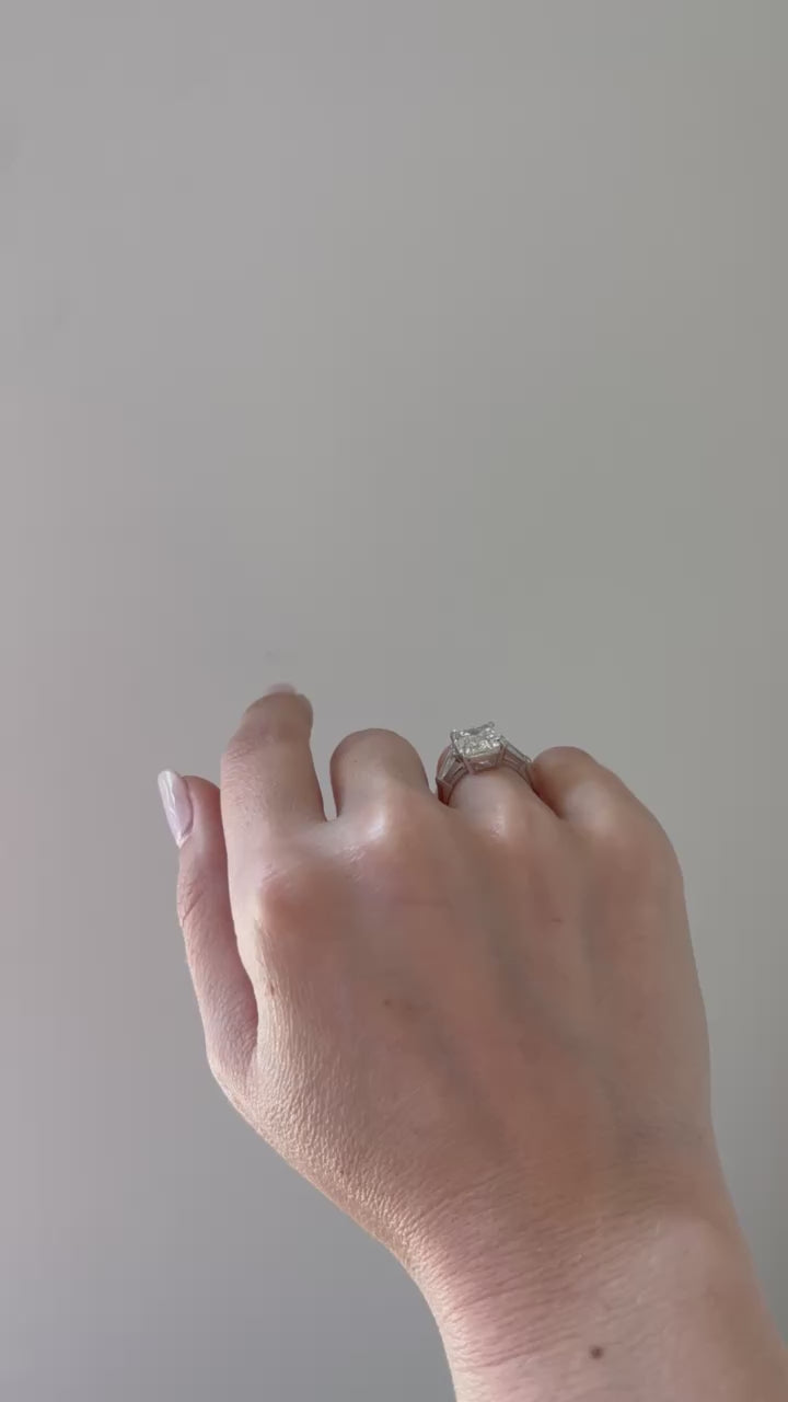 The Lauren Engagement Ring with emerald cut center stone and emerald cut stones on either side with platinum band styled on ring finger of model with no audio