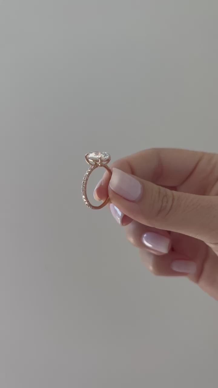 The Jessica Engagement Ring with oval center diamond and micro pave diamonds on rose gold band held in between fingers of model put on ring finger with no audio