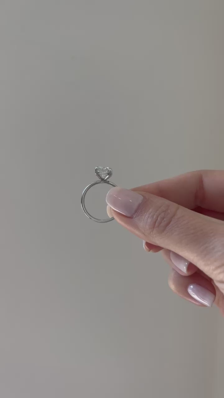 Jamie Engagement Ring with emerald cut center diamond and platinum band held in between fingers of model and put on ring finger with no audio