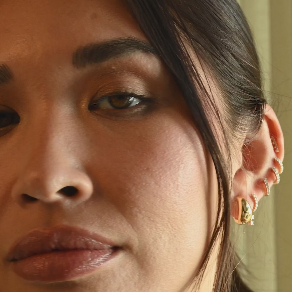 Six diamond and gold earrings styled on ear of model with brown hair with no audio