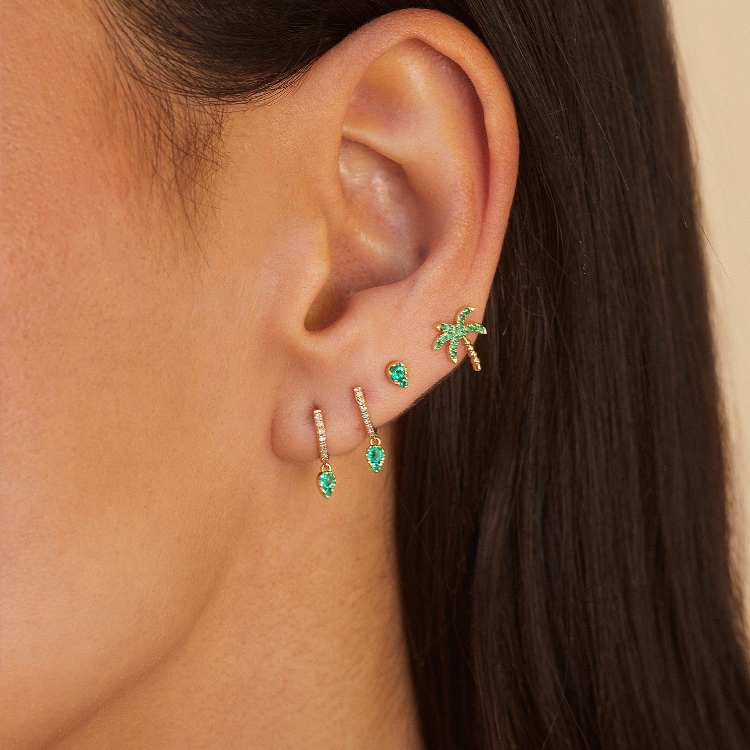 Emerald Teardrop Diamond Mini Huggie Earring in 14k yellow gold styled on first and second earring hole of model next to emerald stud earring and emerald palm tree earring
