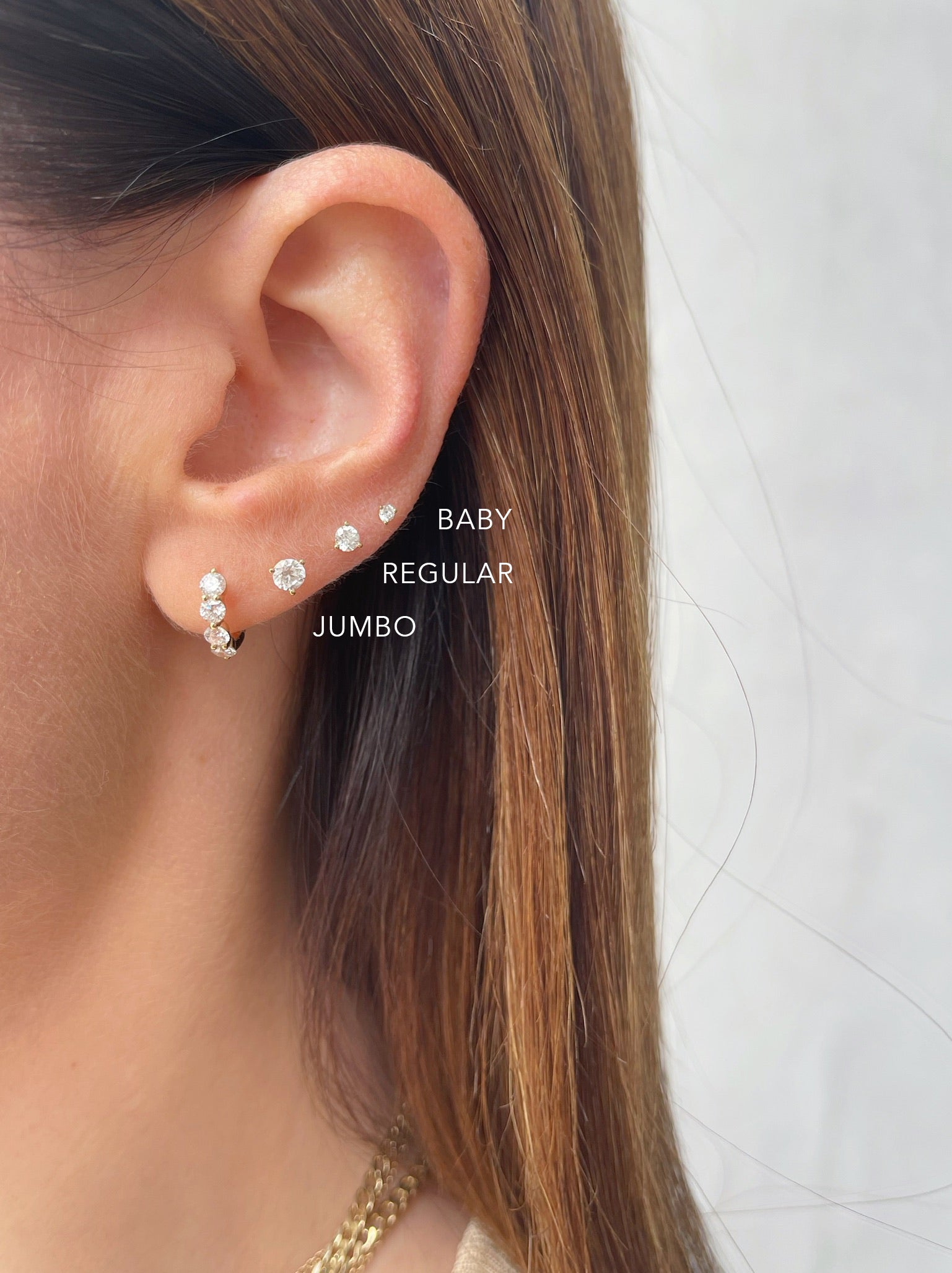 Solitaire Diamond Stud Earring in 14k yellow gold styled on ear with two studs and one huggie earring styled on ear of model diagram with jumbo regular and baby solitaire earring