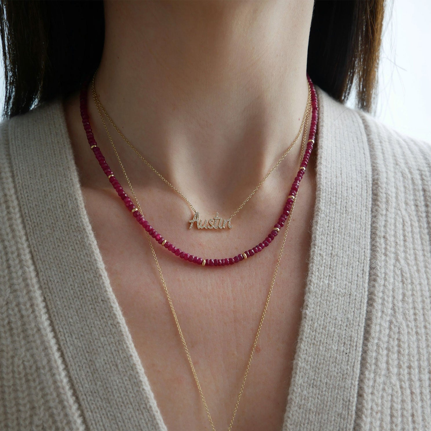 Birthstone Bead Necklace In Ruby styled on neck of model wearing gold and diamond custom script necklace in Austin and wearing tan sweater