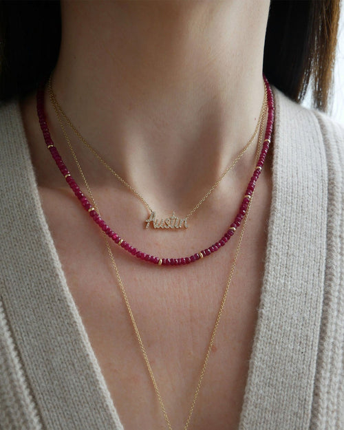 Birthstone Bead Necklace In Ruby styled on neck of model wearing gold and diamond custom script necklace in Austin and wearing tan sweater