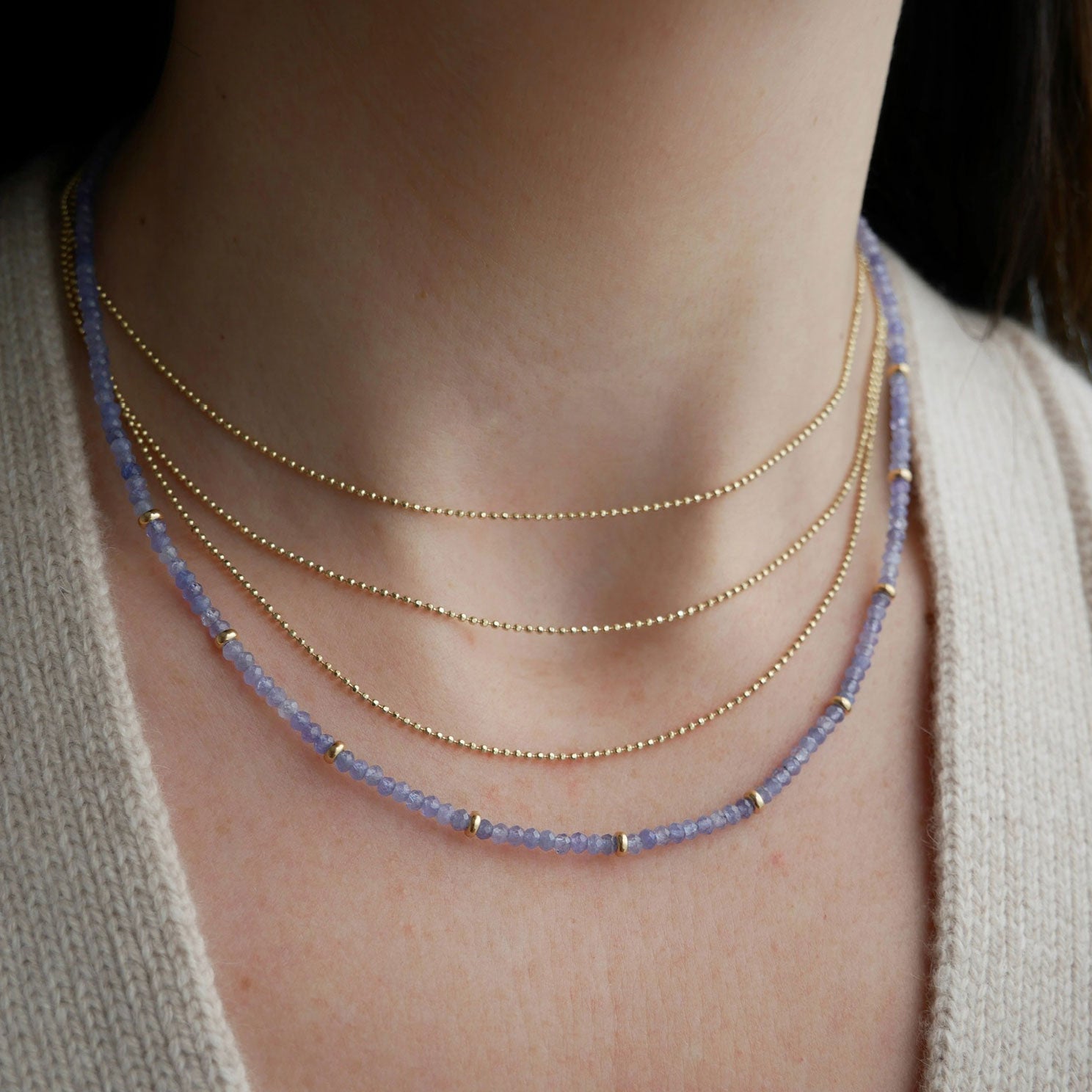 Birthstone Bead Necklace In Tanzanite styled on neck of model with gold layered necklace and wearing tan sweater