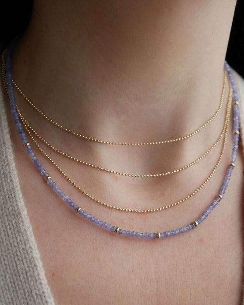 Birthstone Bead Necklace In Tanzanite styled on neck of model with gold layered necklace and wearing tan sweater