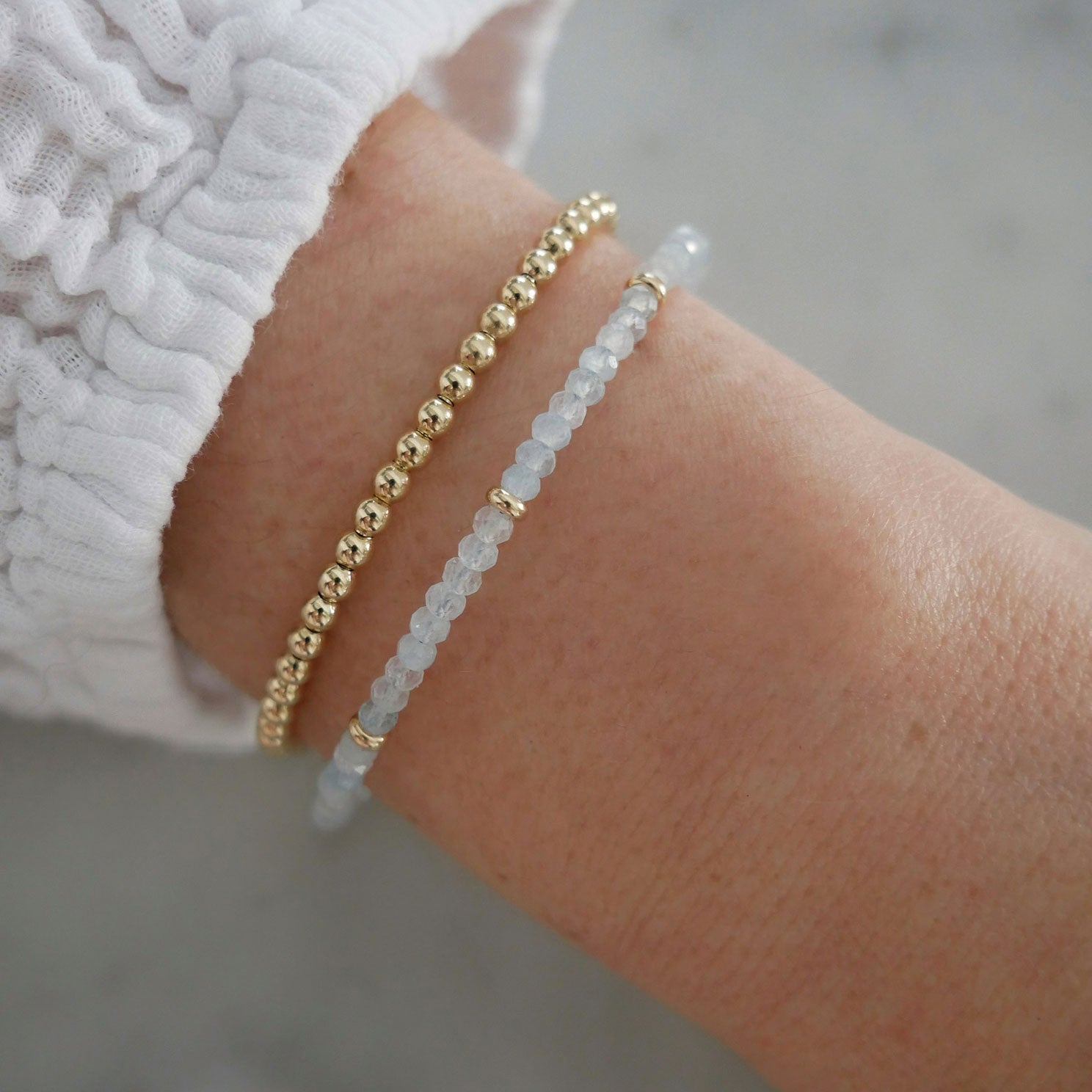 Birthstone Bead Bracelet In Aquamarine styled on wrist of model with gold ball bracelet and wearing white sleeve