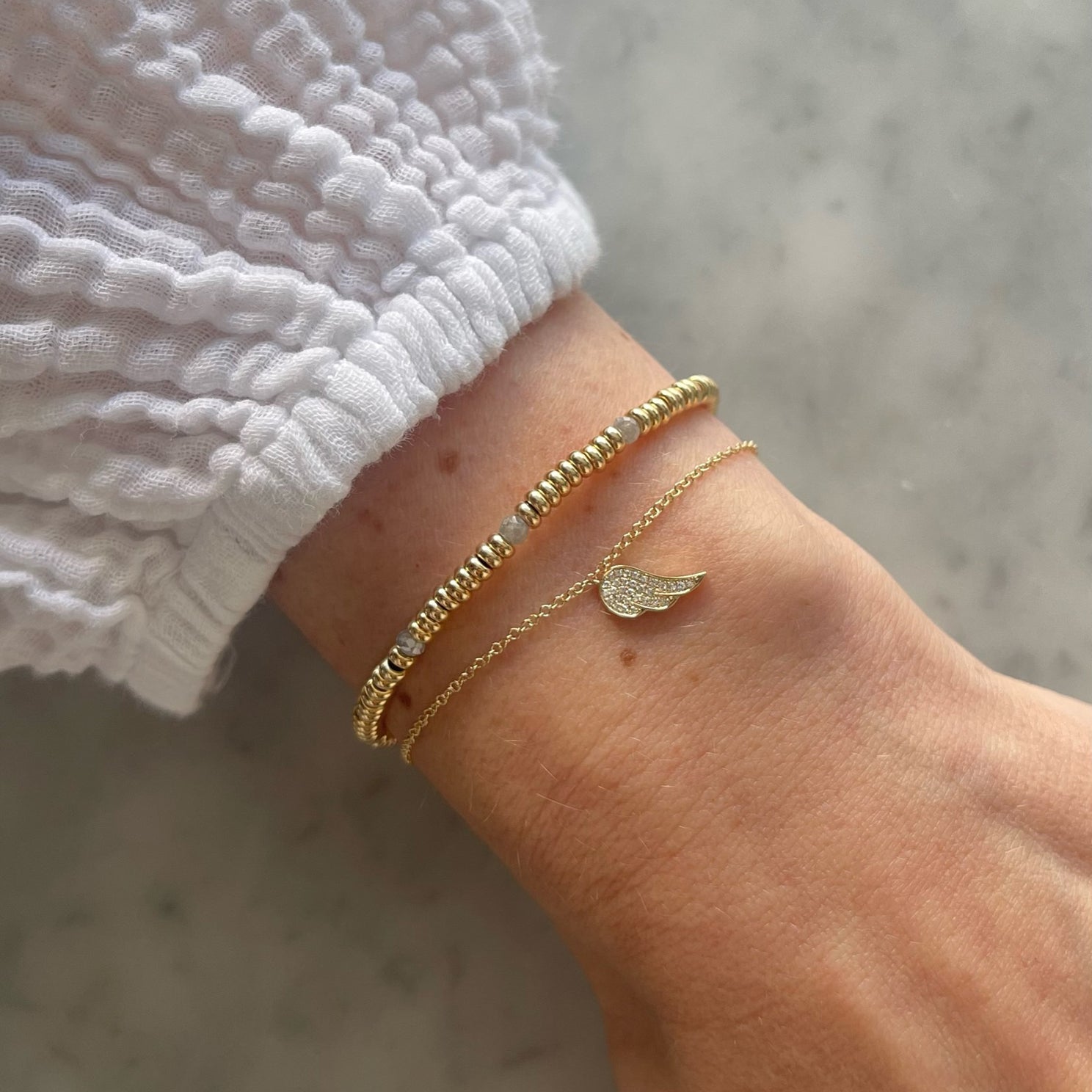 Birthstone Bead Bracelet In Diamond in 14k yellow gold styled next to angel wing bracelet on wrist of model with white sleeve