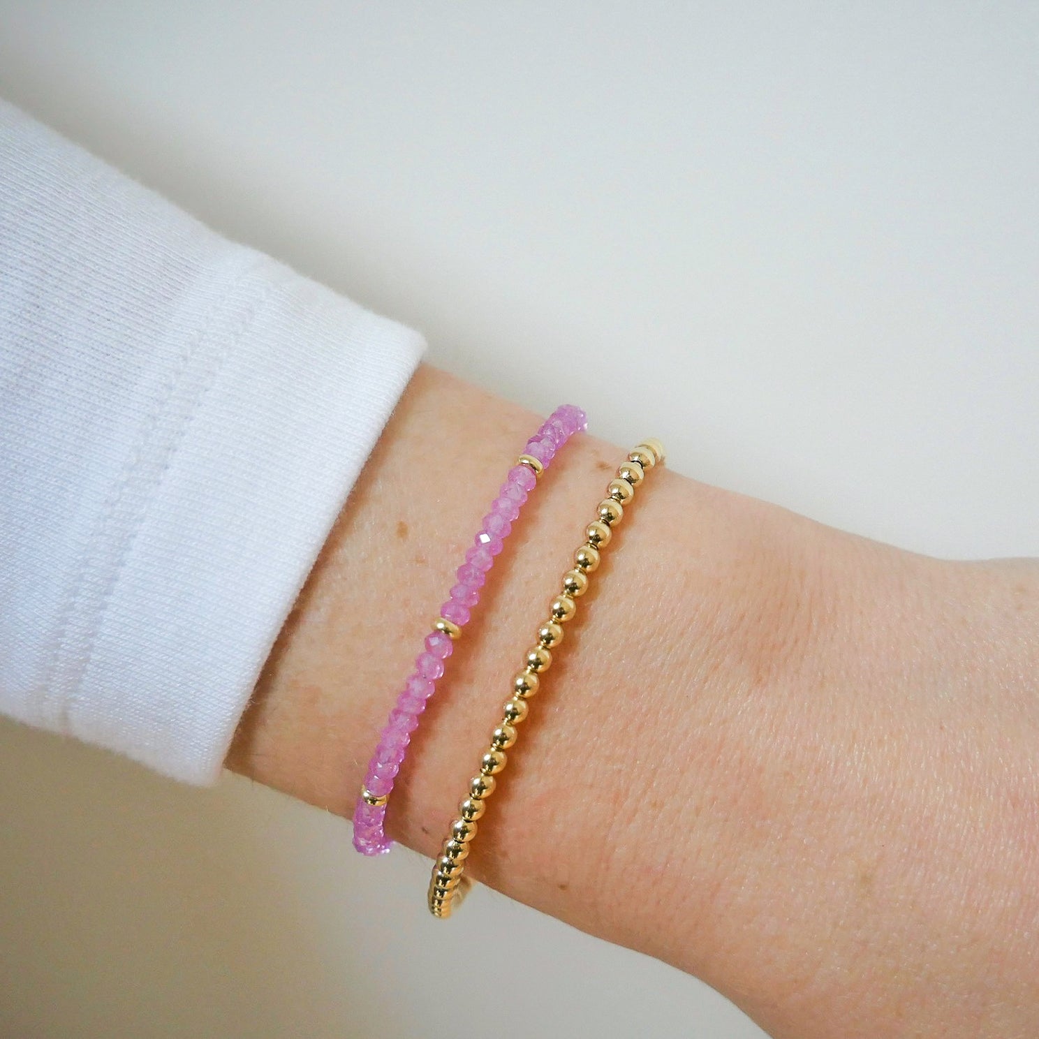 Birthstone Bead Bracelet in Pink Sapphire next to gold ball stretch bracelet styled on wrist of model wearing white sleeve