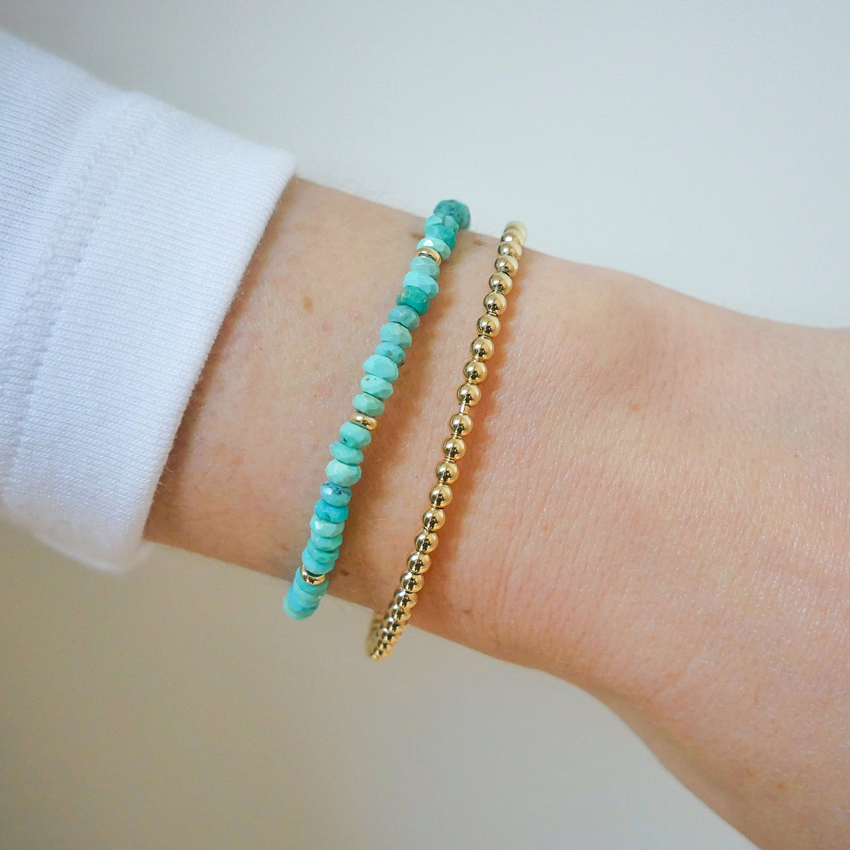Birthstone Bead Bracelet In Turquoise next to gold ball stretch bracelet styled on wrist of model with white sleeve