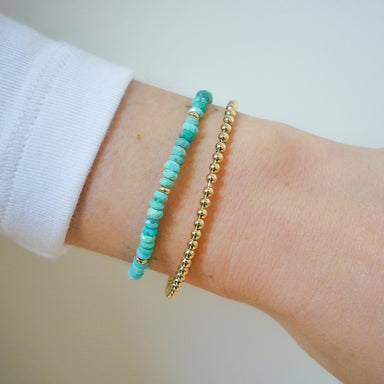 Birthstone Bead Bracelet In Turquoise next to gold ball stretch bracelet styled on wrist of model with white sleeve