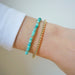 Birthstone Bead Bracelet In Turquoise next to gold ball stretch bracelet styled on wrist of model with white sleeve