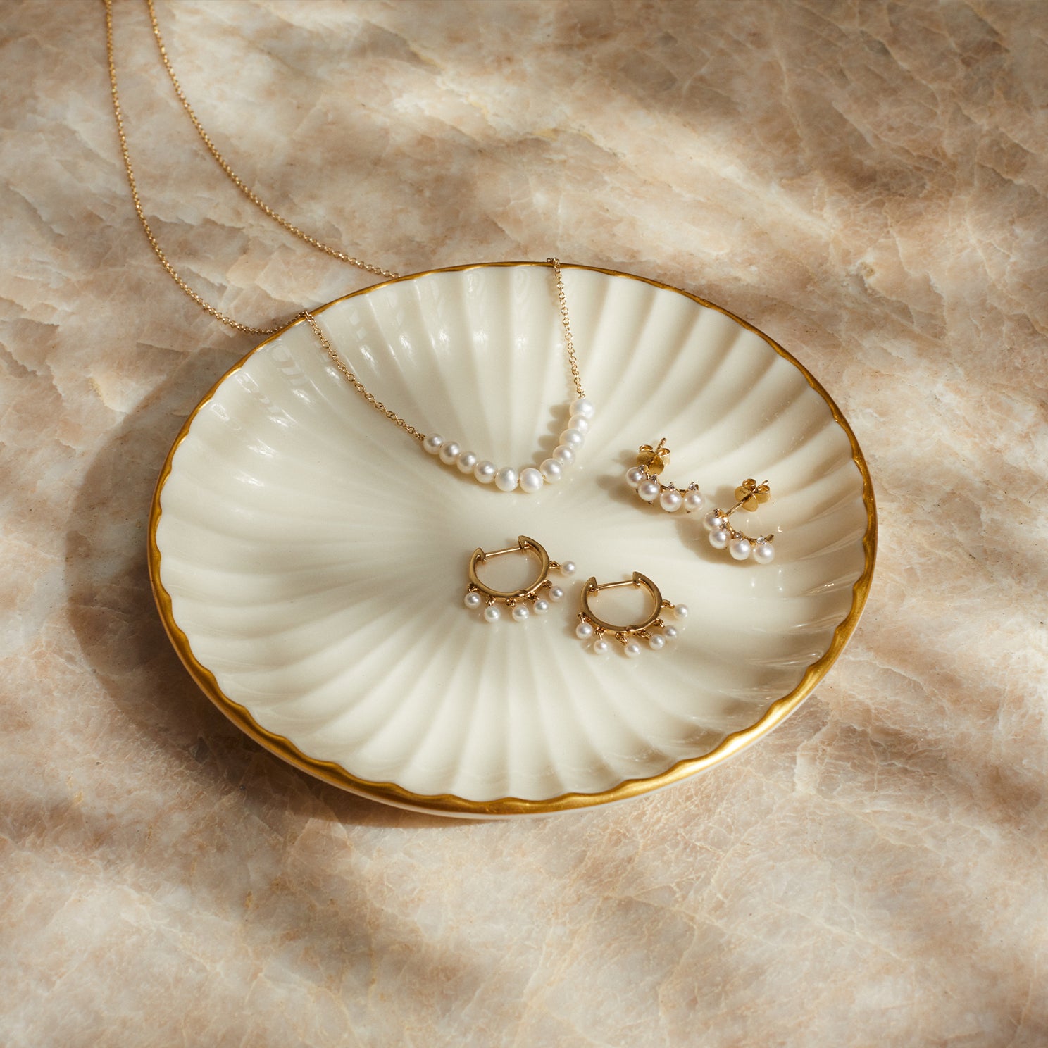 Pearl Necklace in 14k yellow gold laying on top of dish next to mini huggie with pearls and pearl stud earring