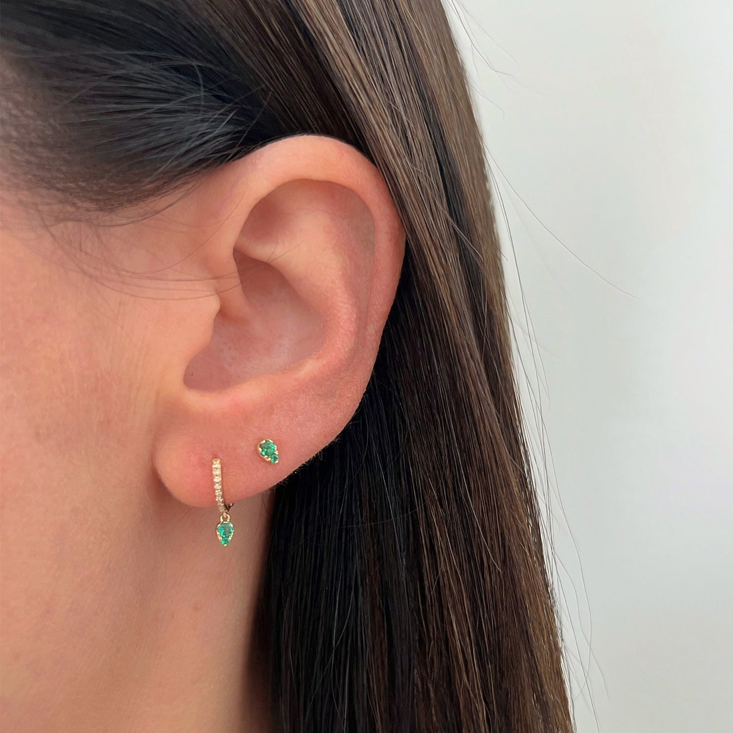 Emerald Teardrop Diamond Mini Huggie Earring in 14k yellow gold next to full cut emerald stud earring both earrings styled on earlobe of model with brown hair