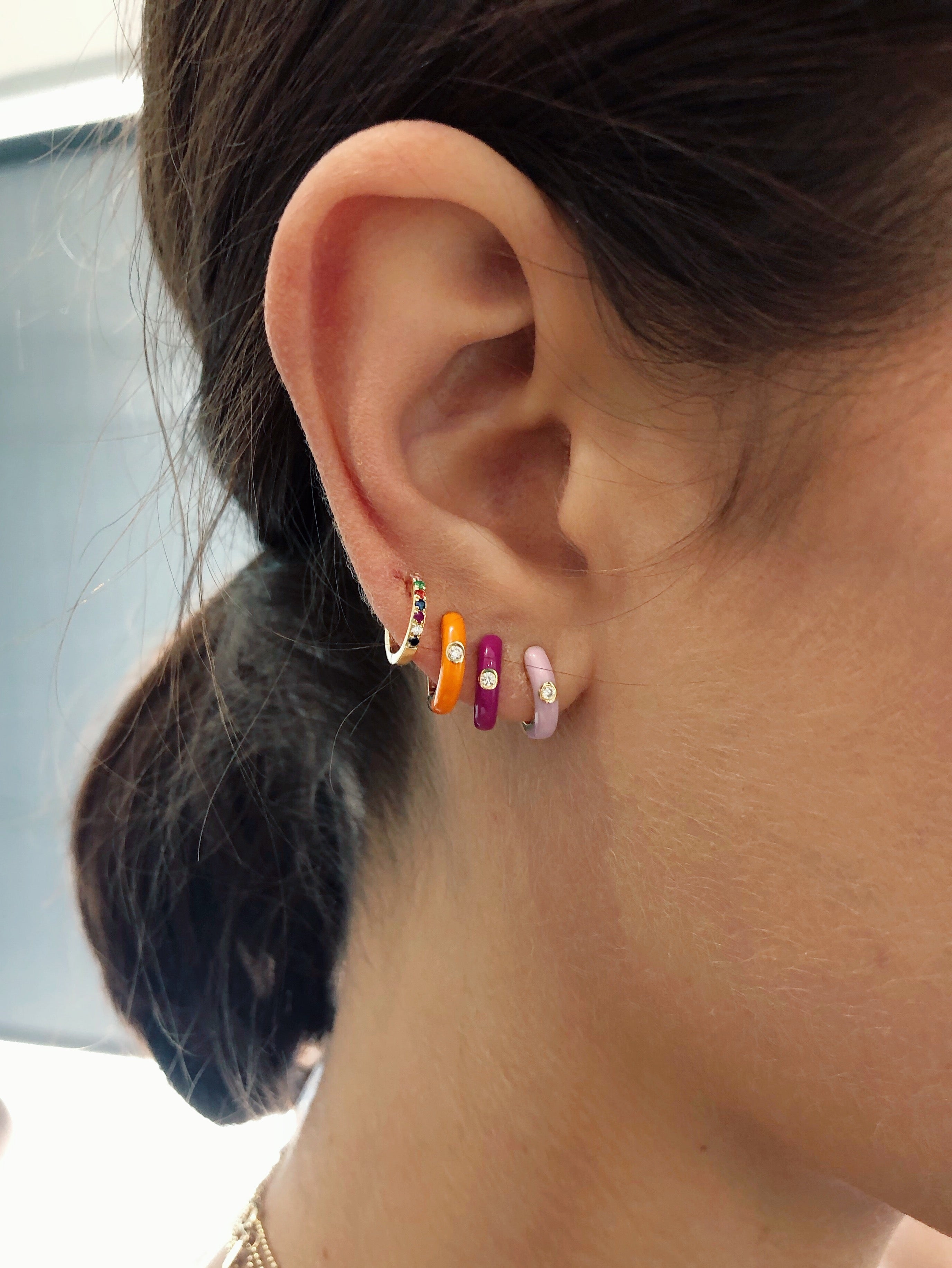 Diamond Orange Enamel Huggie Earring in 14k yellow gold styled on ear lobe of model next to berry enamel huggie, pink enamel huggie, and rainbow huggie