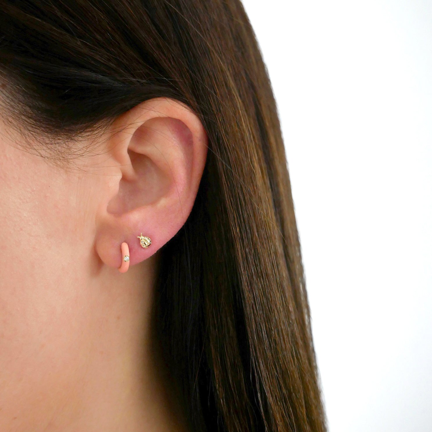 Diamond Coral Enamel Huggie Earring in 14k yellow gold next to gold lady bug stud earring styled on ear lobe of model