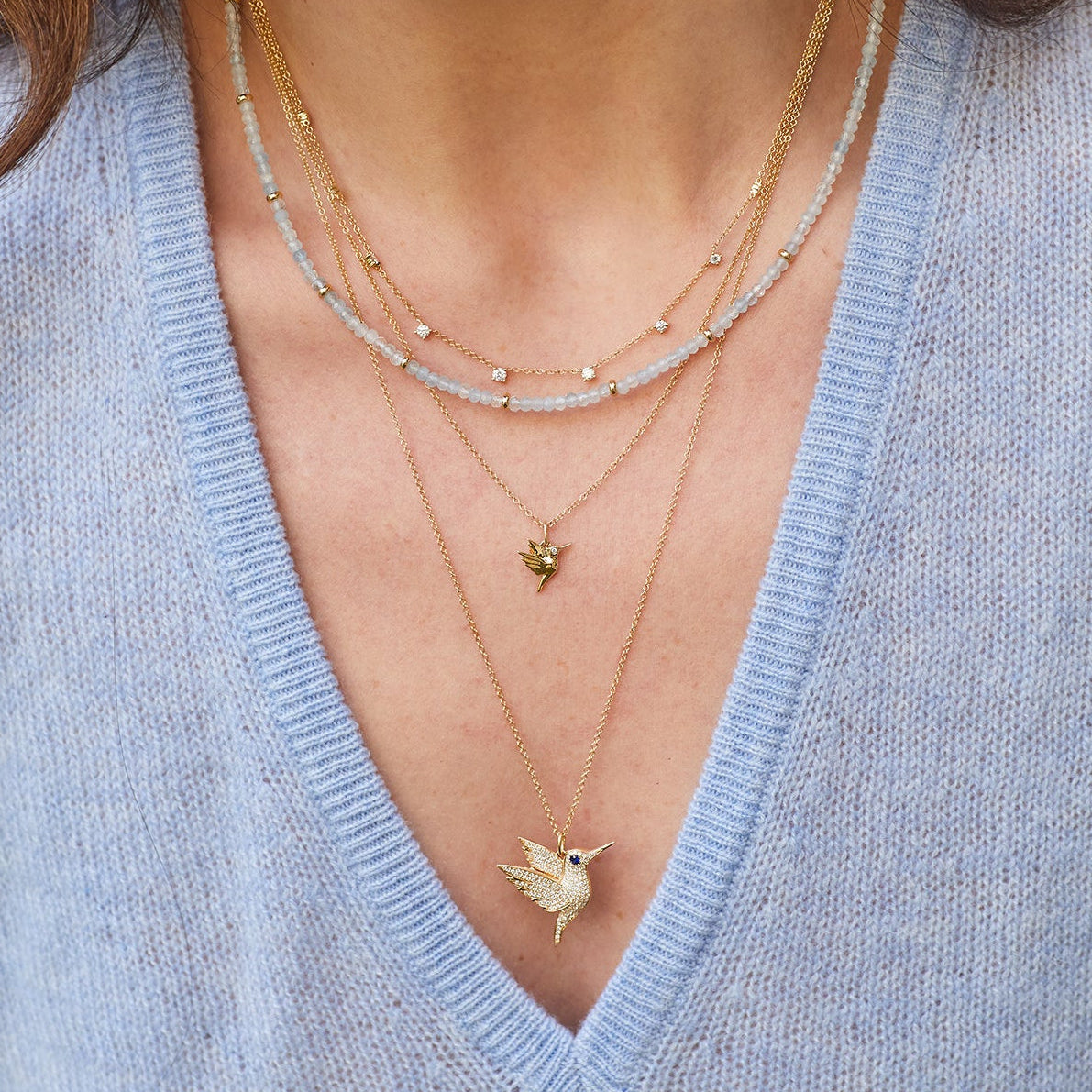 Pavé Diamond Hummingbird Necklace in 14k yellow gold with blue sapphire birthstone eye styled on neck of model layered with three necklaces and wearing blue blouse