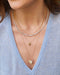 Pavé Diamond Hummingbird Necklace in 14k yellow gold with blue sapphire birthstone eye styled on neck of model layered with three necklaces and wearing blue blouse