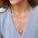 Pavé Diamond Hummingbird Necklace in 14k yellow gold with blue sapphire birthstone eye styled on neck of model layered with three necklaces and wearing blue blouse